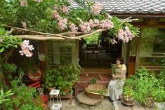 a woman sitting on a wooden bench in front of a house surrounded by plants and flowers