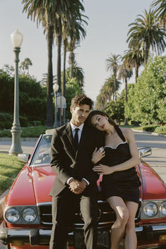 a man and woman sitting on the hood of a red car in front of palm trees