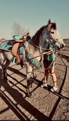 a woman is standing next to a horse in the dirt with a saddle on it's back