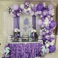 a purple and white dessert table with mermaid balloons on the wall, under an arch