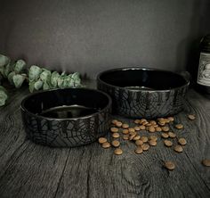 two black pet bowls filled with dog food next to a bottle of wine and leaves