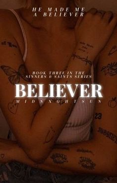 a woman with tattoos on her arms and chest sitting in front of a wall that says believe