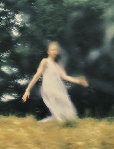 a blurry image of a woman in a white dress walking through the grass with trees behind her