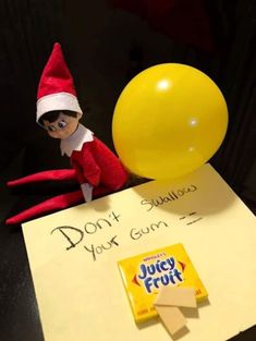 an elf sitting on top of a table next to a yellow balloon and sign that says don't you gum?