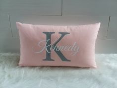 a pink pillow with the letter k in cursive font on it sitting on a fluffy white surface