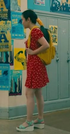 a woman standing in front of a locker with posters on the wall behind her and holding a backpack