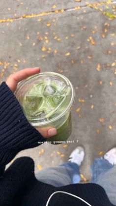 a person holding up a cup filled with green liquid on the street next to some yellow leaves