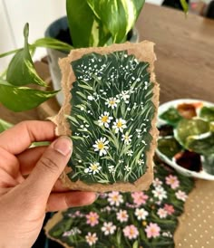 a person holding up a piece of paper with flowers on it and plants in the background