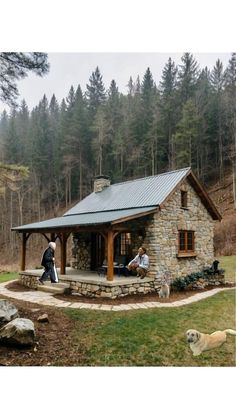 two people sitting on the porch of a small stone cabin with a dog standing next to it