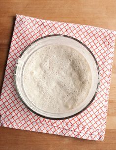 a glass bowl filled with flour sitting on top of a wooden table next to a red and white checkered cloth