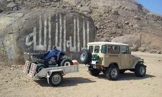 a jeep towing a motorcycle on a trailer in front of a large rock with writing written on it