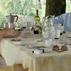 two people sitting at a table with wine glasses and plates on it, in front of trees