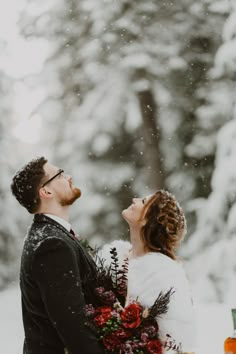 a bride and groom standing in the snow with their arms around each other looking up