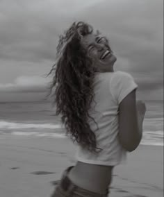 a woman standing on top of a beach next to the ocean with her hair blowing in the wind