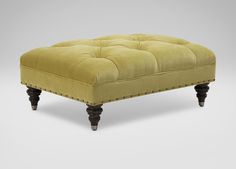 an ottoman with wooden legs and a buttoned tufted cushion on the top, sitting in front of a gray background