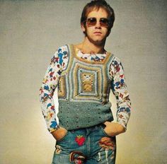 a man with sunglasses on standing in front of a wall wearing jeans and a sweater