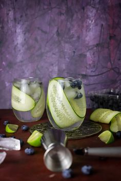 two glasses filled with cucumber and blueberries on top of a wooden table