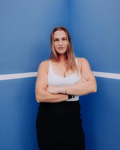 a woman standing in front of a blue wall with her arms crossed and looking at the camera