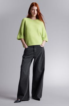 a woman standing in front of a gray background wearing black pants and a lime sweater