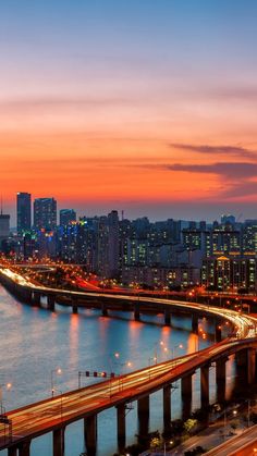 the city skyline is lit up at night, with cars driving on the road and bridge in the foreground