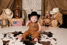 a baby sitting on a cow print rug wearing a cowboy hat and brown overalls