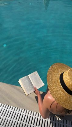 a woman sitting on the edge of a swimming pool reading a book while wearing a straw hat