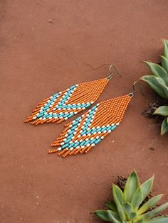 an orange and blue beaded earring laying on the ground next to succulents