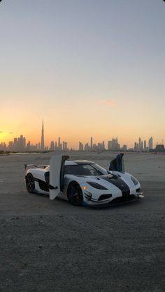 a white and black sports car parked in the middle of a desert area with a city skyline behind it