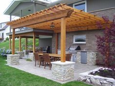 an outdoor kitchen and dining area is shown in this backyard setting with stone walls, wood pergol