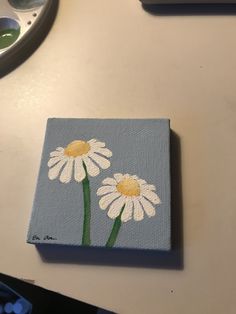 two white daisies painted on a blue square with yellow centers sitting on a table