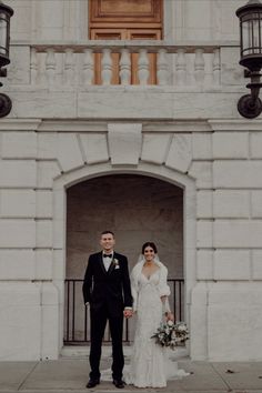 a bride and groom standing in front of a white building with an arched doorway holding hands