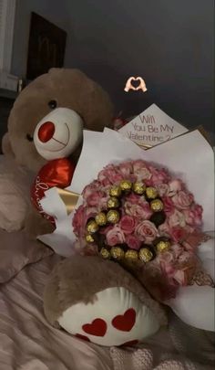 a teddy bear sitting on top of a bed holding a bouquet of roses and chocolates