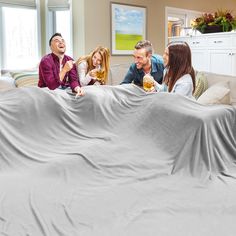 four people sitting on a couch with a blanket covering them and drinking beer in the living room