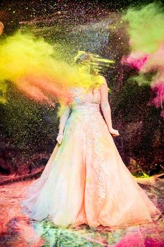 a woman standing in front of colored powder on her face and dress, with arms outstretched