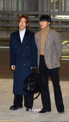 two men standing next to each other in front of an airport terminal, one wearing a coat and the other carrying a black handbag