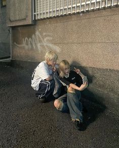 two people sitting on the ground next to a wall with graffiti written on it and one person talking on a cell phone