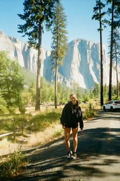 a woman is walking down the road in front of some trees and mountains with a car