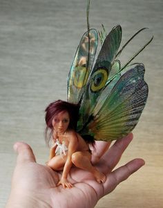 a tiny doll sitting on top of a hand with a butterfly wing attached to it