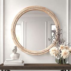 a round mirror on the wall above a table with flowers in vases and books