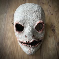 a white mask with black eyes and teeth on a wooden surface, that looks like it has been made out of paper machs