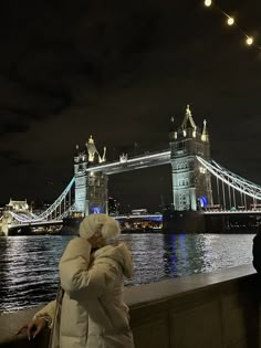 a woman standing on the side of a river next to a tall bridge at night