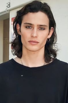 a woman with long dark hair wearing a black shirt and silver hoop earrings, standing in front of a white wall