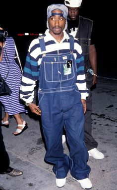 a man wearing overalls and a hat standing next to another man on the street