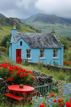 a blue house surrounded by wildflowers in the mountains with a red table and chair