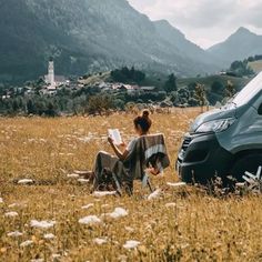 a woman sitting on the ground next to a van in a field with mountains behind her