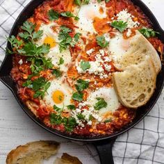 an iron skillet filled with eggs and tomato sauce next to bread on a white table