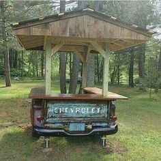 an old truck is parked in the grass under a wooden structure with a picnic table attached to it