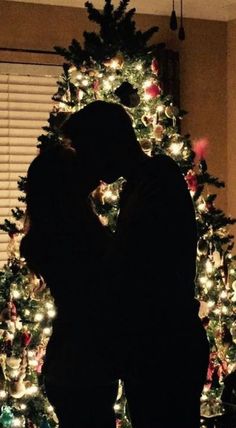 a man and woman kissing in front of a christmas tree with lights all around it