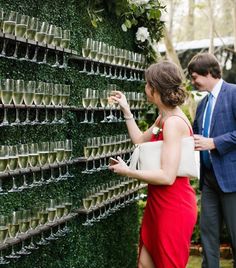 a man and woman standing next to a wall covered in cups with wine glasses on them