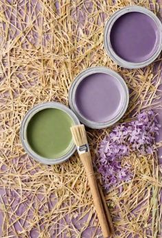 three paint cans with different shades of purple and green sitting on straw next to a brush